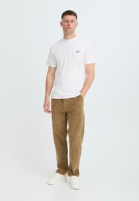 White short-sleeve t-shirt with a small black logo, paired with brown corduroy pants and white sneakers. Model stands against a light background.