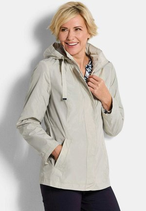 Lachende vrouw met lichtbeige gewatteerde jas met capuchon, hand in de zak, kraag rechtzettend, staand tegen een effen witte achtergrond.