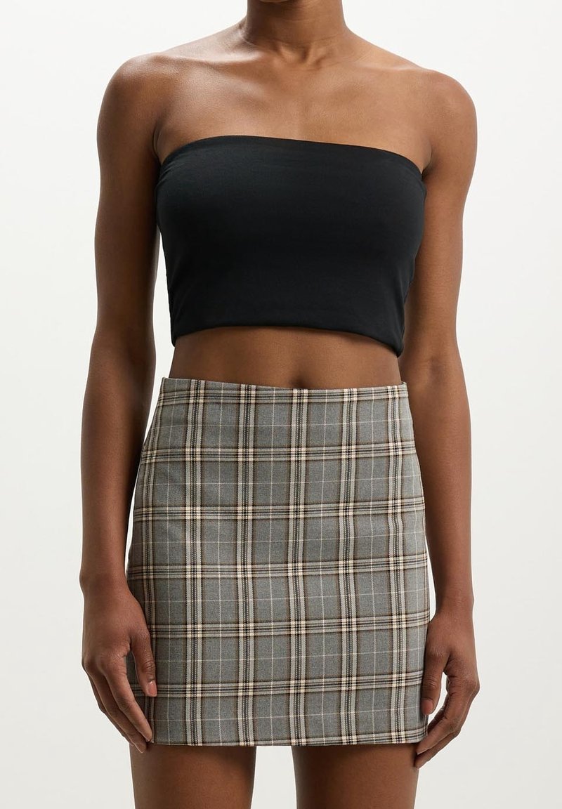 Femme portant un crop top noir sans bretelles et une mini-jupe grise à carreaux taille haute, debout devant un fond uni.