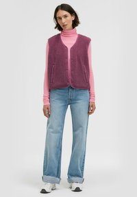 Fleeceweste in Pflaume mit strukturierter Oberfläche, offener Vorderseite und zwei Seitentaschen. Getragen über einem rosa Rollkragenpullover, kombiniert mit hellblauen Jeans.