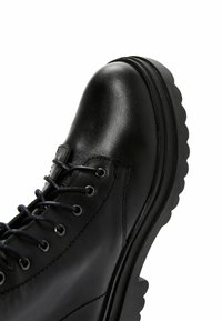 Stivaletto in pelle nera con punta tonda, design con lacci, superficie testurizzata e suola spessa e rigata. Presenta occhielli e cuciture in argento.
