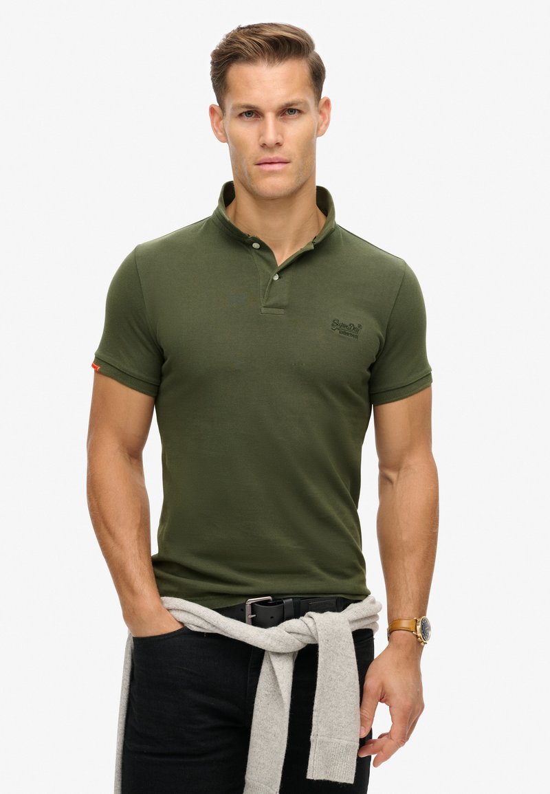 Camiseta polo verde oliva con un tejido texturizado, placket de tres botones y mangas cortas. Suéter gris claro atado a la cintura.