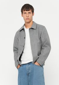 Grijs geruite jas met een kraag, knoopsluiting aan de voorkant en zijzakken, gecombineerd met een wit shirt en lichtblauwe jeans.