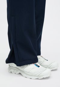 Ναυτικές φόρμες jogger με λεπτομέρειες από φερμουάρ στο πλάι, συνδυασμένες με λευκά αθλητικά παπούτσια με γκρι λεπτομέρειες και πολύχρωμο λογότυπο στη γλώσσα.