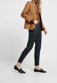 Blazer marron ajusté sur un col roulé marron, associé à un pantalon slim noir à motifs et des chaussures plates noires. Design texturé et ajusté.