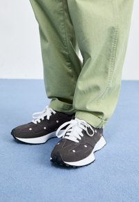 Dunkelgraue Sneakers mit floraler Stickerei und weißen Schnürsenkeln, kombiniert mit hellgrünen Baumwollhosen, auf einem blauen Teppich.