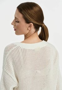 Biały sweter z dzianiny o luźnym, otwartym splotcie, z szerokim dekoltem i opadającymi szwami ramion. Teksturowany materiał podkreśla miękkość.