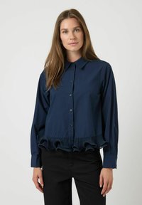 Vrouw in een marineblauwe knoopblouse met lange mouwen en een geplooide volantzoom, gecombineerd met zwarte broek.