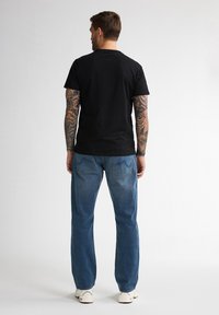 Zwarte katoenen t-shirt, blauwe denim jeans, zichtbare achterzakken, gedragen met witte sneakers. Model gezien vanaf de achterkant tegen een lichte achtergrond.
