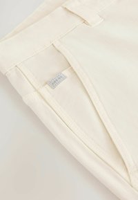Cream-colored cotton trousers featuring a smooth texture, multiple stitching details, and a visible brand tag near the waistband.