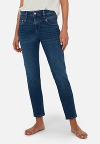 Jeans de mezclilla azul oscuro con corte slim, cintura de media altura, cinco bolsillos y un ligero desgastado. Los dobladillos están remangados y se pueden ver acentos de metal.