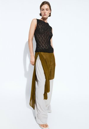 Donna con top nero senza maniche in pizzo, pantaloni larghi grigio chiaro e una lunga gonna a portafoglio verde oliva, in piedi davanti a uno sfondo bianco.