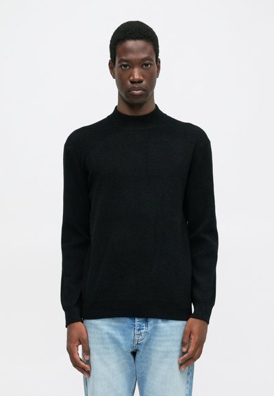 Giovane uomo che indossa un maglione nero a collo alto e jeans azzurri chiari, in piedi contro uno sfondo bianco semplice.