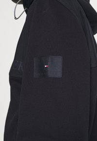 Sudadera negra con capucha, fabricada en un tejido texturizado y con un parche con el logo de Tommy Hilfiger cosido en la manga izquierda.
