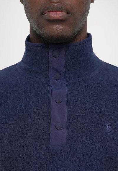 Marineblå fleece-pullover med høj krave, prydet med knaplukning og en diskret logo-detalje. Blød tekstur og ensartet farve hele vejen igennem.