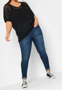 Svart öppenstickad topp med korta ärmar, ihop med åtsittande blå denimjeans. Jeanssen har en lätt blekning och upprullade nederkanter. Svarta sneakers med vita sulor.