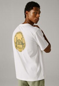 Junger Mann blickt über die Schulter, trägt ein weißes T-Shirt mit gelbem Vintage-Grafik des Brighton Piers auf dem Rücken, steht vor neutralem Hintergrund.