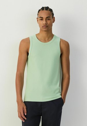 Junger Mann mit geflochtenen Haaren trägt ein hellgrünes ärmelloses Tanktop und dunkle Hosen, steht vor einem einfachen hellen Hintergrund.