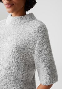 Maglione grigio chiaro realizzato in tessuto morbido e testurizzato. Presenta un alto colletto e maniche corte, con una superficie morbida e pelosa.