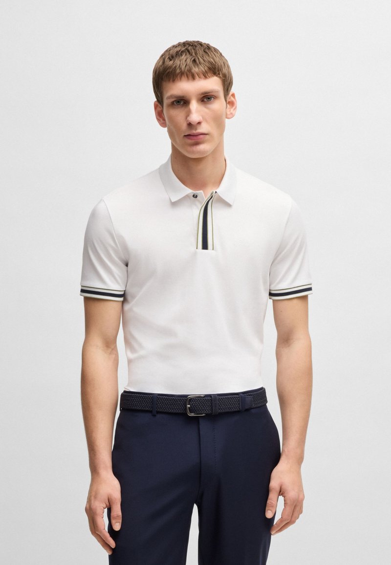 Biała koszulka polo z krótkim rękawem, z dwoma guzikami przy dekolcie, w granatowe i żółte paski na kołnierzyku i rękawach, połączona z granatowymi spodniami.