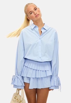 Chemise bleu clair à manches longues et à boutons, avec des poignets plissés, associée à une jupe courte à volants assortie. Tissu texturé, coupe décontractée.