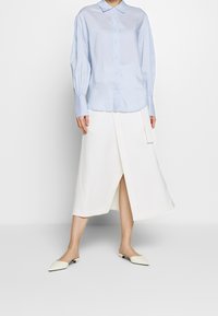 Camicia button-up azzurro chiaro con colletto e maniche lunghe; abbinata a una gonna midi asimmetrica bianca e scarpe slip-on color crema.
