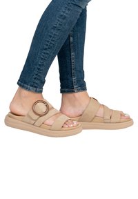 Des sandales à glissière beiges avec de larges sangles en suède, un accent métallique circulaire et une semelle en caoutchouc rembourrée. Portées avec un jean bleu foncé.