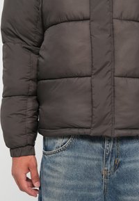 Polstret mørkegrå jakke med quiltet design, der har en stående krave og elastiske manchetter, parret med lyseblå denimjeans.