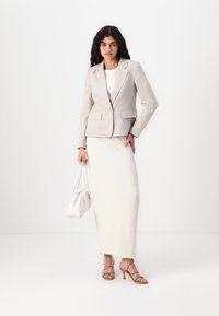 Sacou tailored de culoare bej deschis, cu două buzunare în față, asociat cu o fustă maxi albă texturată și sandale cu toc subțire de culoare bej, ținând o geantă mică albă.