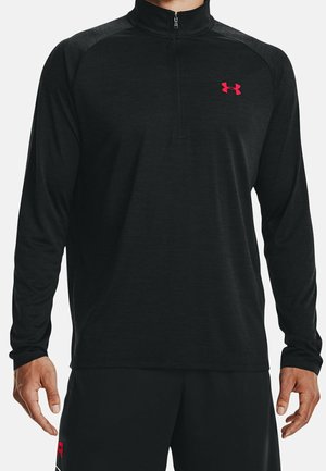 Mann trägt ein schwarzes, langärmliges Sportshirt mit Viertel-Reißverschluss und rotem Under Armour-Logo auf der Brust sowie schwarze Shorts mit roten Details.