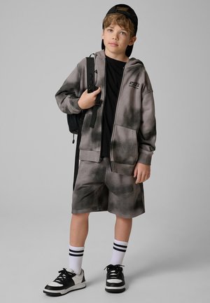 Chłopiec noszący czarną czapkę do tyłu, szarą bluzę tie-dye, pasujące szorty, czarną koszulkę, białe skarpetki w paski, czarno-białe trampki i czarny plecak.