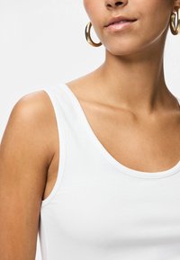 Κοντινό πλάνο μιας γυναίκας που φοράει λευκό αμάνικο πάνω μέρος και μεγάλα χρυσά κρίκους στα αυτιά, δείχνοντας το λαιμό, τον ώμο και το κάτω μέρος του προσώπου της.