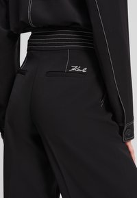 Black trousers with a fitted waistband, featuring white stitching and embroidered logo. Smooth fabric with side pockets and button accents.