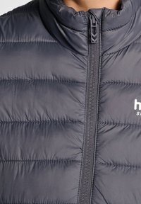 Grå pufferväst med hög krage, med horisontell sömnad, en framdragkedja och en liten logotyp på vänster bröstområde.