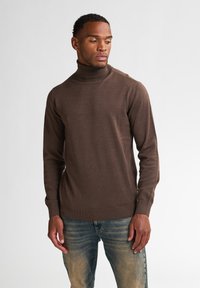 Pull à col roulé marron en matériau doux et à maille fine. Présente des manches longues et des poignets et un ourlet côtelés ajustés. Porté avec un jean bleu clair.