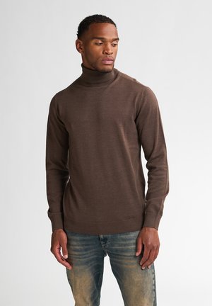 Suéter marrón de cuello alto en un material suave de punto fino. Presenta mangas largas y puños y bajo de canalé ajustados. Combinado con jeans de color azul claro.