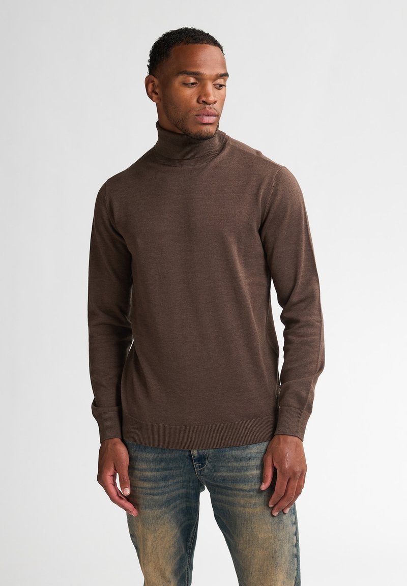 Pull à col roulé marron en matériau doux et à maille fine. Présente des manches longues et des poignets et un ourlet côtelés ajustés. Porté avec un jean bleu clair.