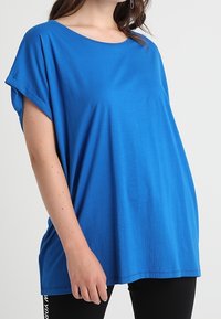 Žena v pohodlném jasně modrém tričku s krátkými rukávy s ohrnutými lemy a černých legínách na jednobarevném světlém pozadí.