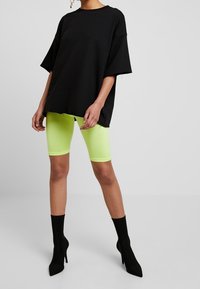 T-shirt nera oversize a maniche corte, abbinata a leggings verde neon. Stivaletti neri con punta affilata. Sfondo bianco semplice.