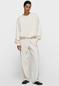 Sweatshirt blanc oversize avec poignets côtelés, associé à un pantalon évasé assorti avec taille à cordon de serrage et détails de couture latérale. Chaussures noires.