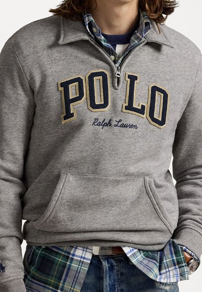 Szürke polár pulóver félcipzárral, nagy "PO LO" felirattal és "Ralph Lauren" hímzéssel. Kék és zöld kockás ing fölé rétegezve.