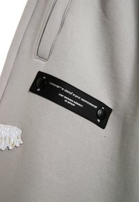 Tela gris con textura; presenta una etiqueta negra con texto blanco y un botón. Se puede ver un diseño bordado en blanco con detalles dorados.