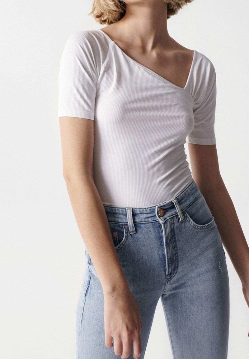 Body bianco a costine con design a spalla sola, maniche corte e silhouette aderente, abbinato a jeans azzurri a vita alta.