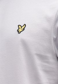 T-shirt de algodão cinza claro com gola redonda e logótipo de águia preto e dourado bordado na área do peito esquerdo. Textura suave.