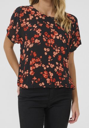 Kobieta ubrana w czarną bluzkę z krótkim rękawem w czerwono-różowy wzór kwiatowy, w połączeniu z czarnymi spodniami, stoi z jedną ręką w kieszeni.