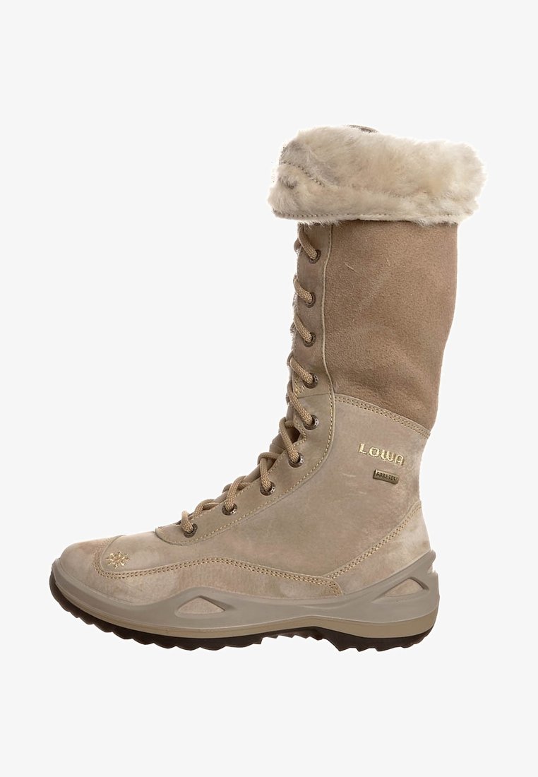 Knæhøj støvle i lysbrun ruskind med en kunstpelskant, snørelukning foran og tekstureret gummisål. Har et guldlogo på siden.