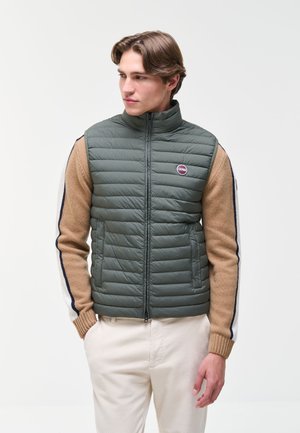 Junger Mann trägt eine grüne gesteppte Weste über einem beigen Pullover mit gestreiften Ärmeln und hellen Hosen und schaut nach links.