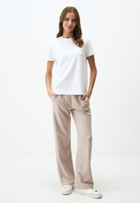 T-shirt bianco a maniche corte, vestibilità rilassata, abbinato a pantaloni beige a gamba larga in tessuto morbido. Modella in piedi con le mani in tasca, indossa sneakers bianche.
