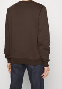 Felpa marrone con scollo rotondo, polsini e orlo a costine, e un tessuto liscio e strutturato. La vista posteriore mostra un design minimalista e senza loghi visibili.
