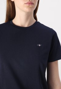 Tamsiai mėlynas medvilnės marškinėliai su apvaliu iškirpimu. Kairėje krūtinės pusėje yra mažas siuvinėtas logotipas. Trumpi rankovės.